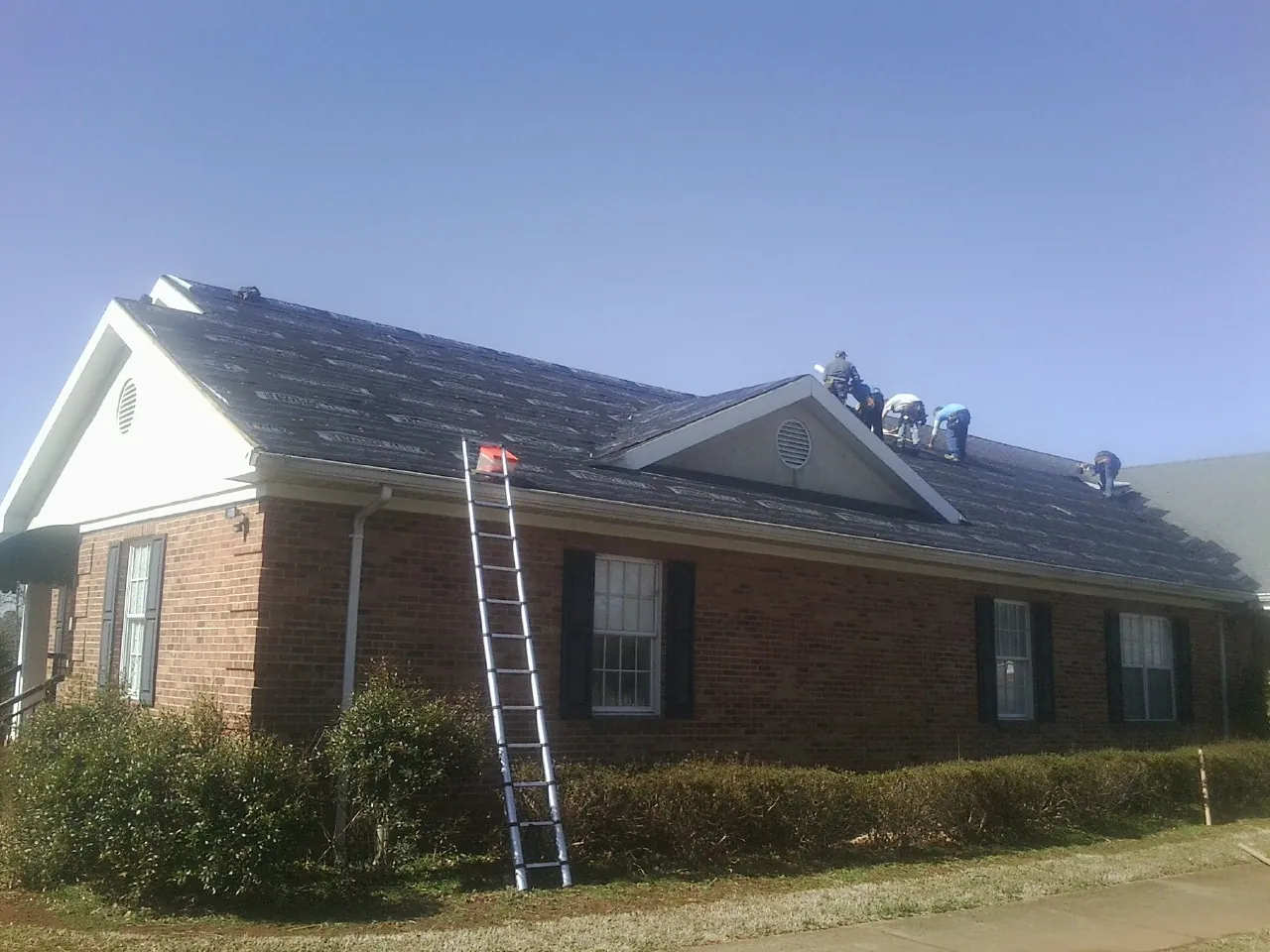 Crew tearing shingles off a brick ranch roof with a ladder set — Emergency Roof Repair work by our crew in West Babylon