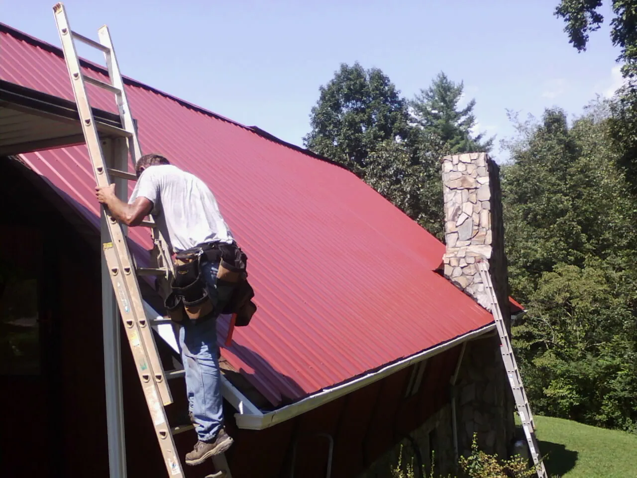 Roofer on a ladder beside a red metal roof and stone chimney — Soffit & Fascia Repair by Roofing Brothers in West Babylon