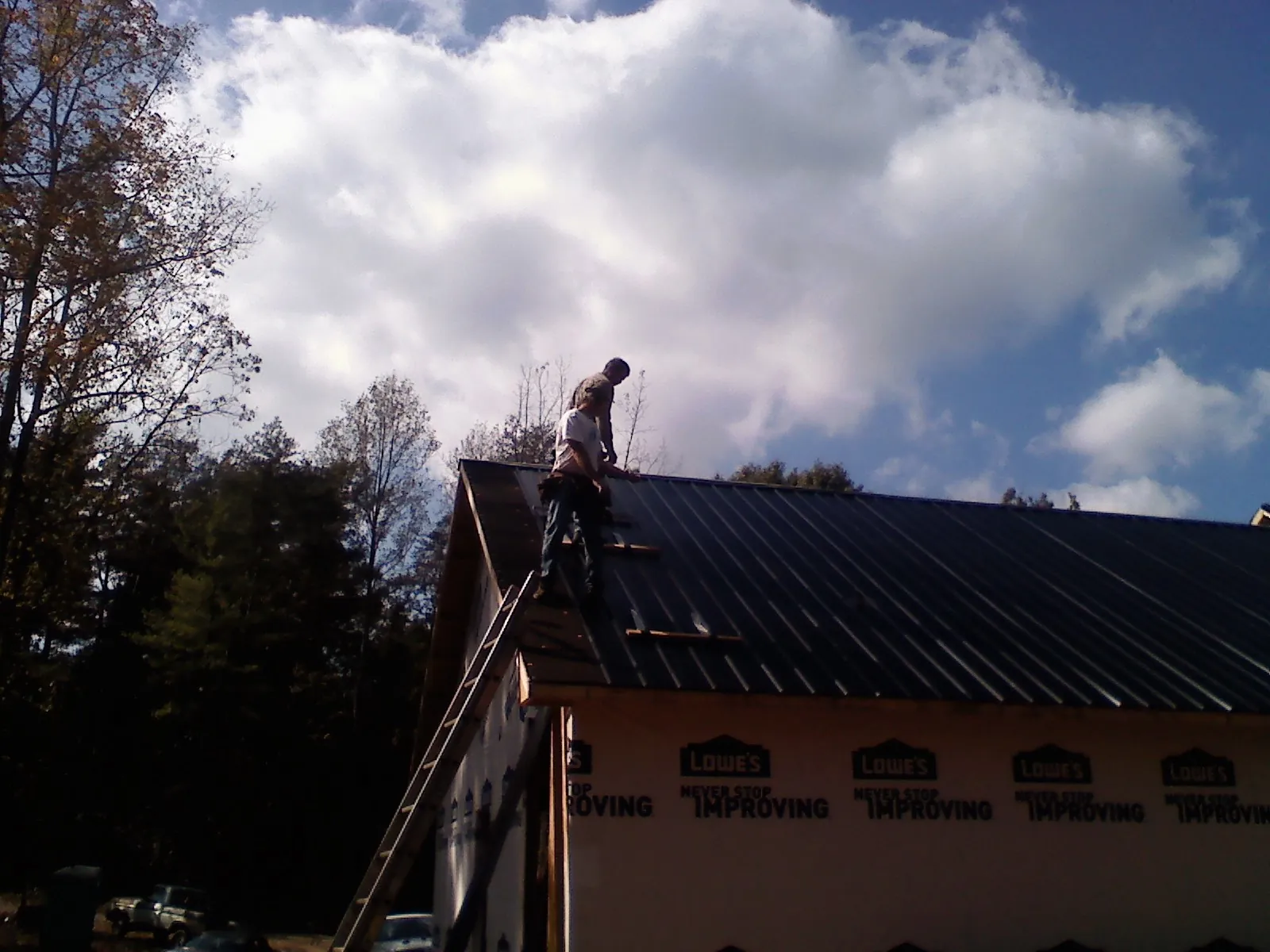 Roofer fastening dark metal panels high against a cloudy sky, from a Roof Inspection job in West Babylon